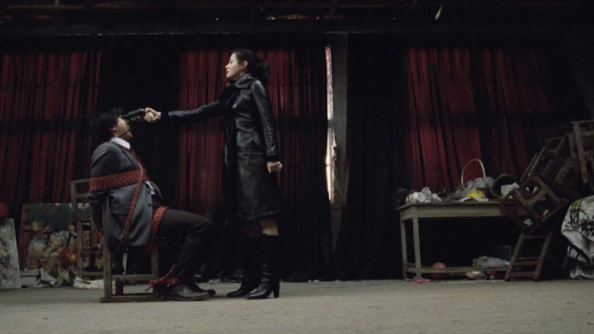 woman wearing black outfit pointing gun to the head of man sitting in char