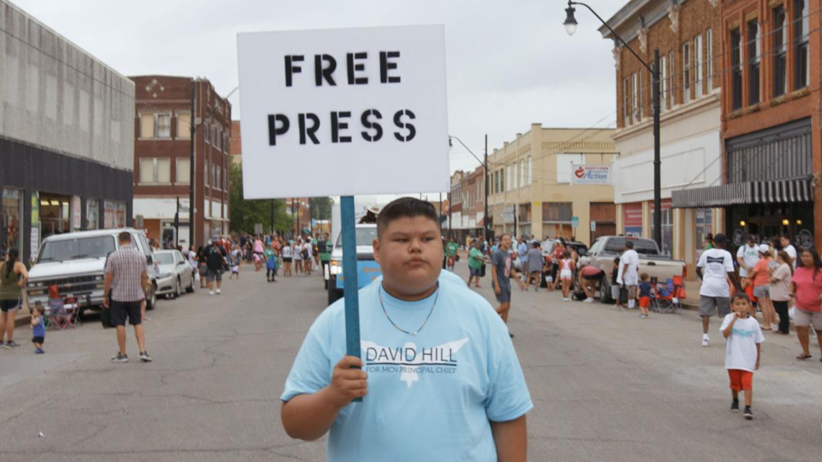 young teen in blue shirt standing in middle of street holding sign that says "Bad Press"