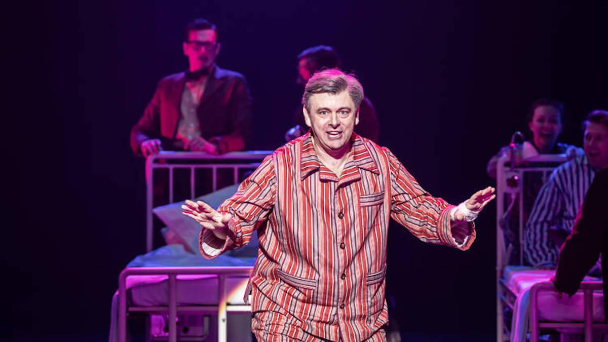 man standing on stage in striped pajamas with single beds in background