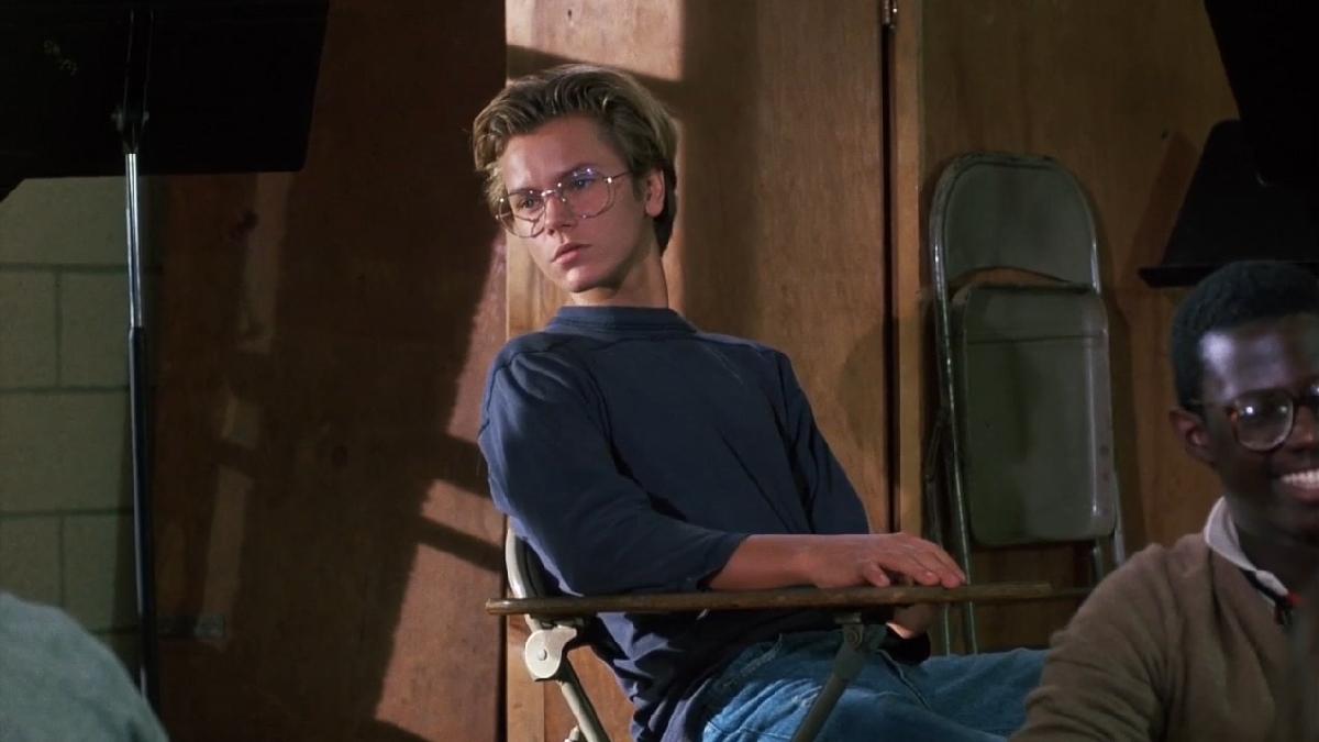 student wearing blue sweater and glasses seated at desk leaning back and looking in distance