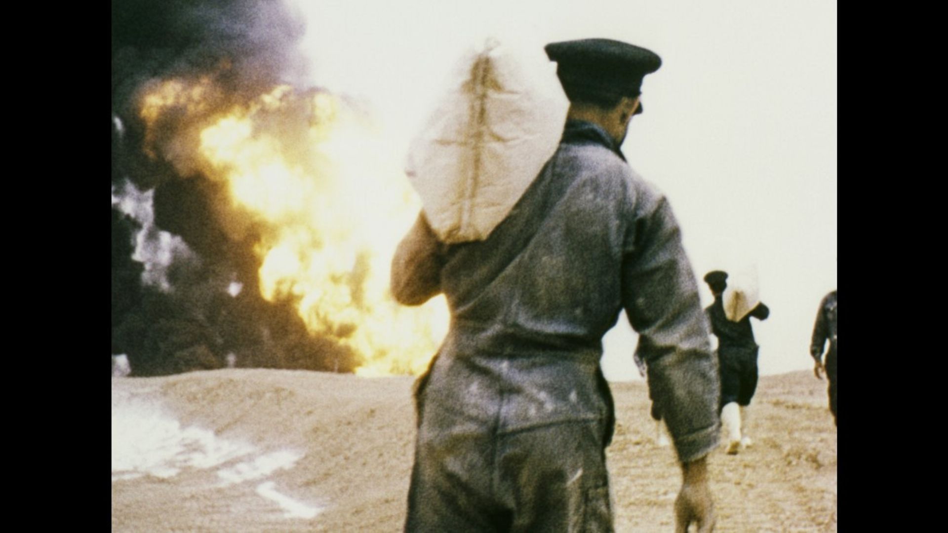 man in military uniform with bag over shoulder walking towards large smoke and fire