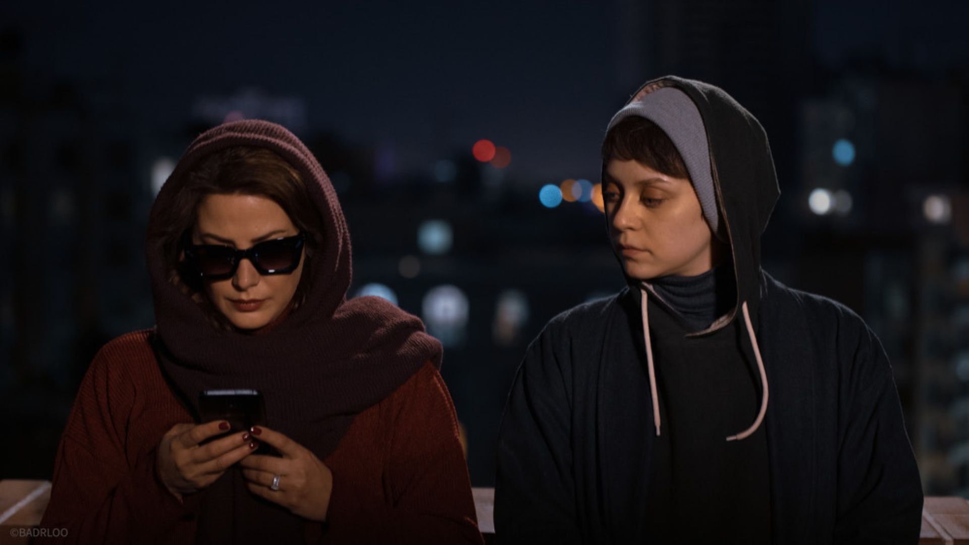 two woman with hoods up on street with one woman wearing sunglasses looking down at phone