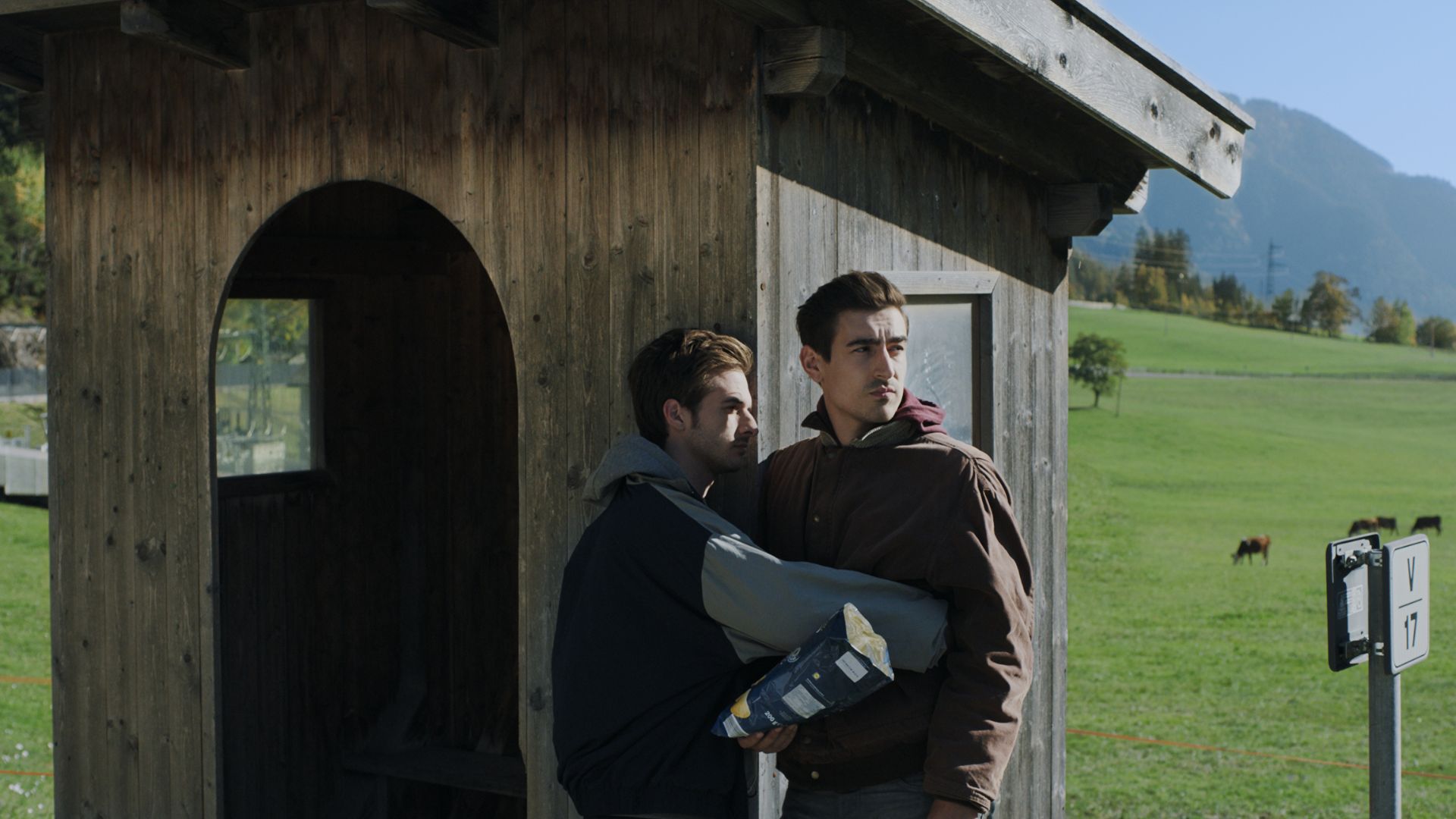 THE MAN WITH THE ANSWERS, 2021, two men standing in front of wooden cabin looking out with fields and mountains in background