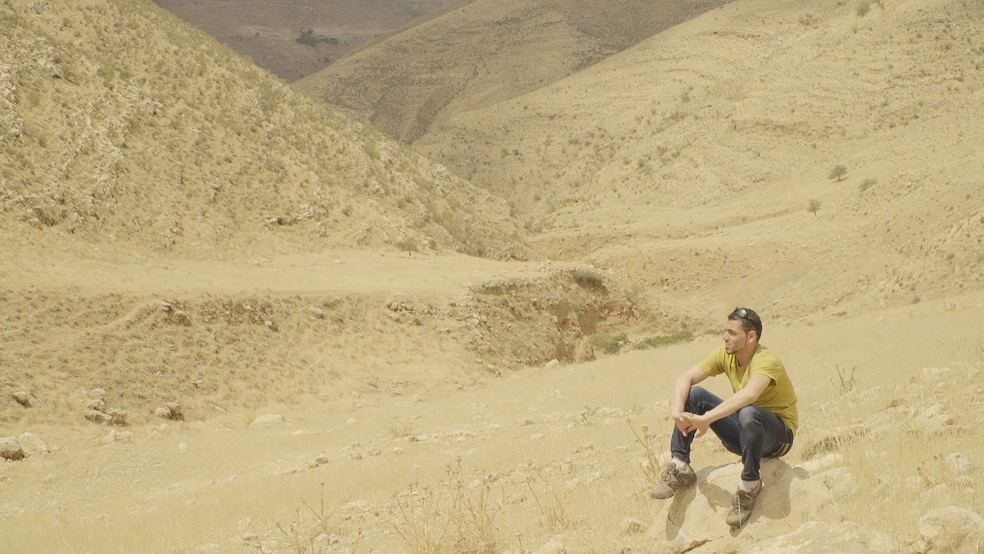 HOLIDAYS IN PALESTINE, 2024, man sitting in vast desert on ground with arms resting on his legs