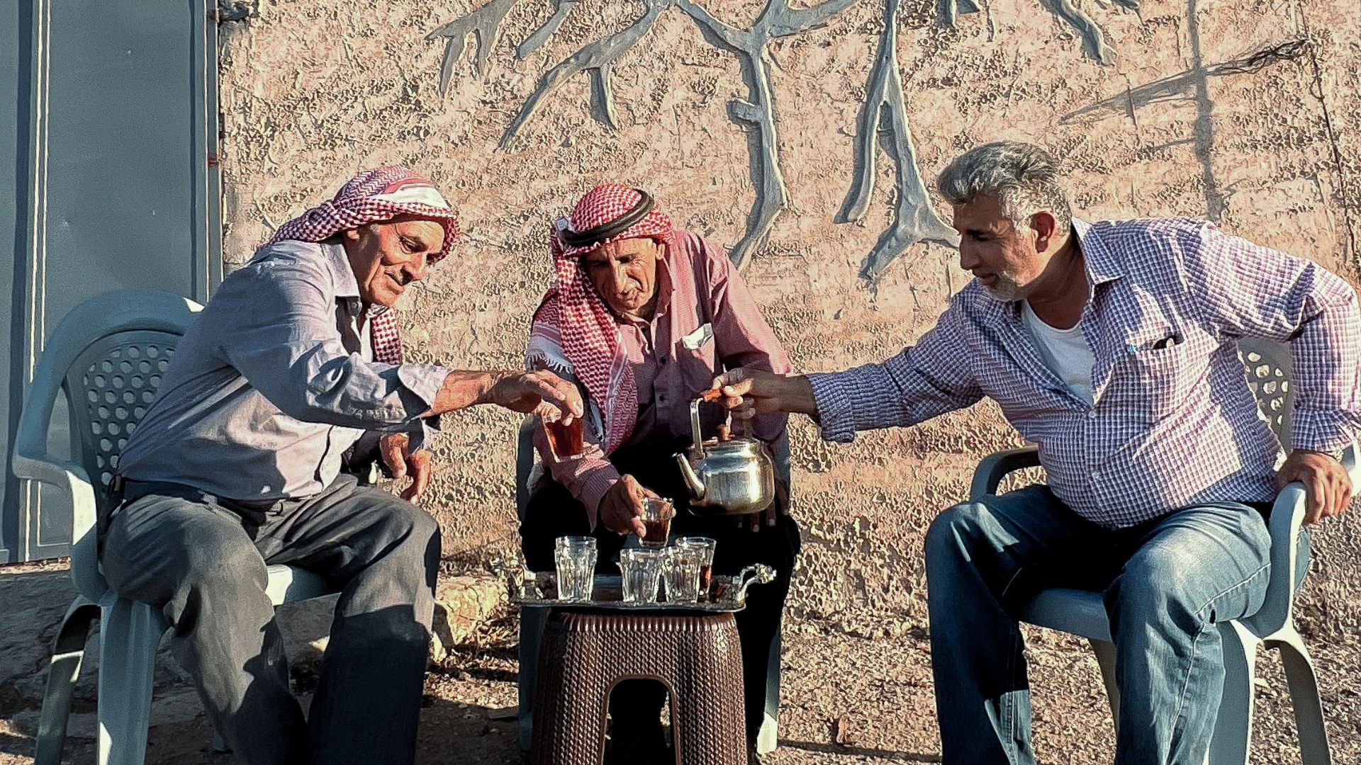 LAND OF CANAAN, 2025, three men seated on plastic charis outside around a small table cheersing drinks together