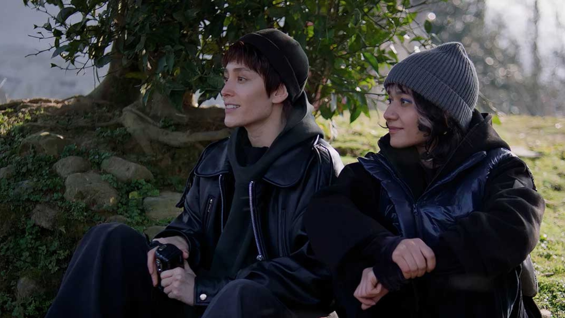 BETWEEN DREAMS AND HOPE, 2025, two young people seated outside wearing coats and arms crossed