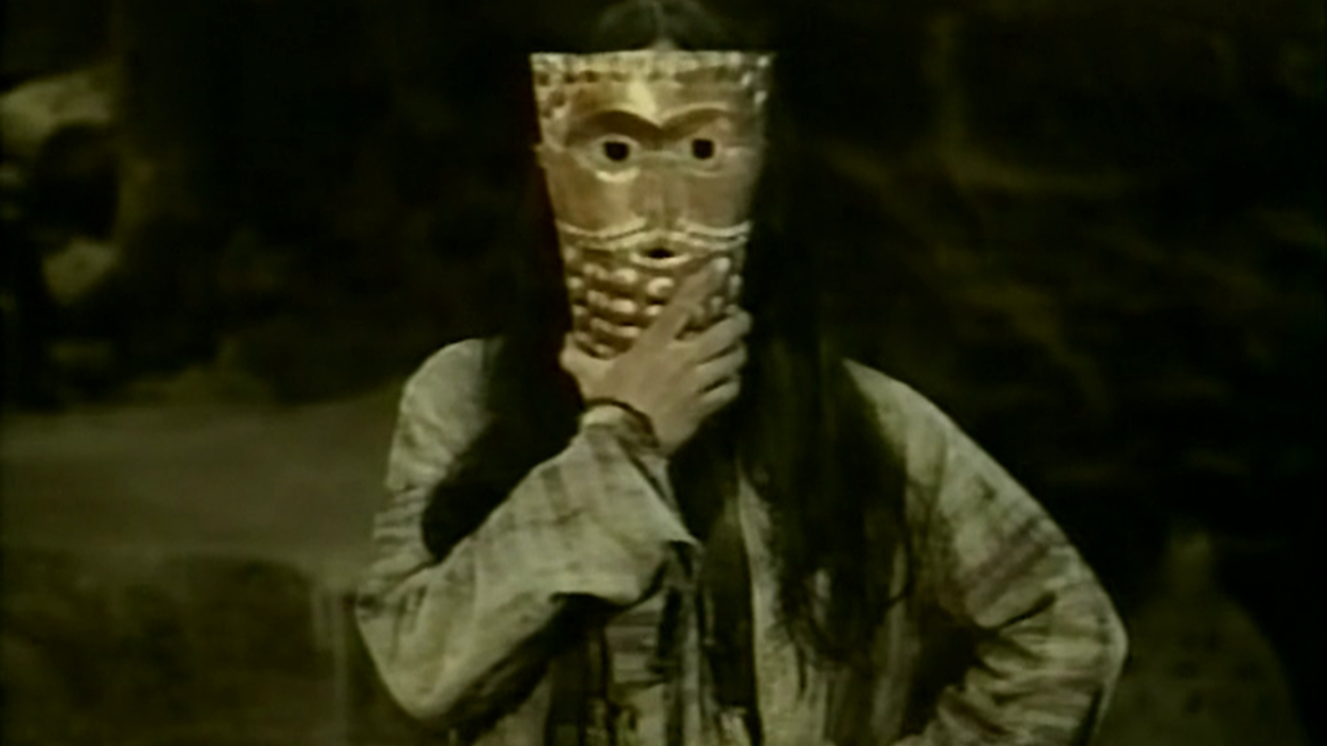 THE DEATH OF YAZDGERD, 1982, person with long hair wearing long sleeves holding up gold mask over face