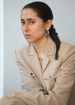 A Moroccan woman with dark black hair pulled back in a tight braid wearing a khaki colored jacket and hoop earrings looking directly at the camera. 
