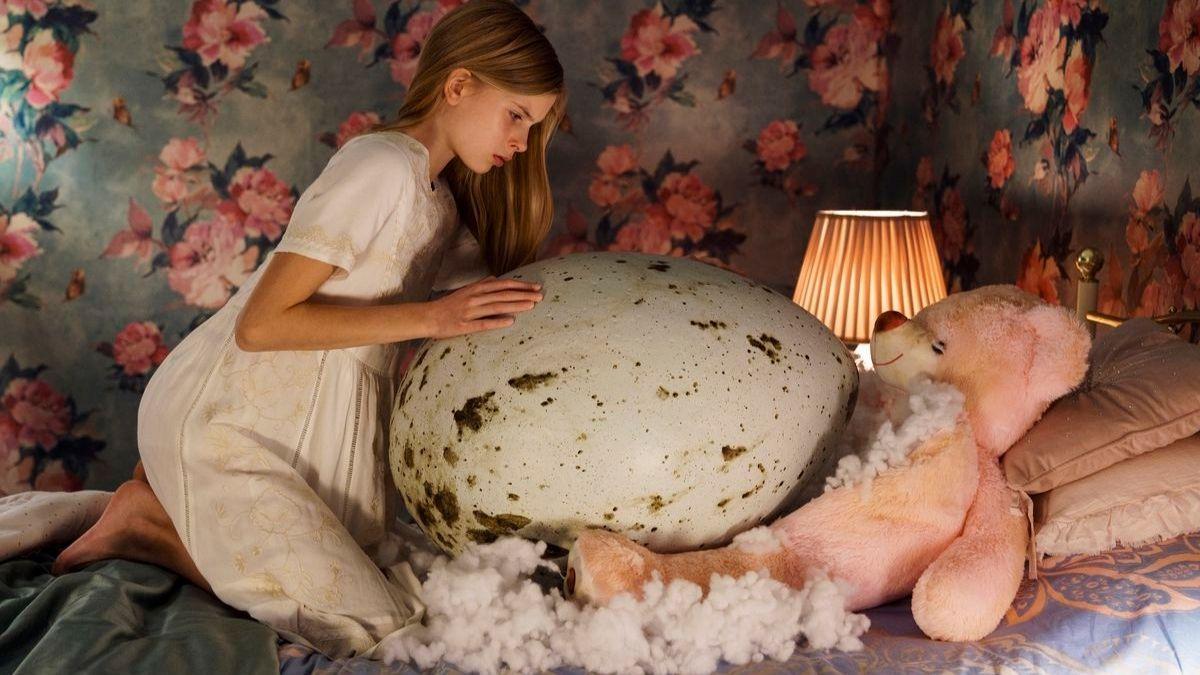 girl standing over large egg with teddy bear