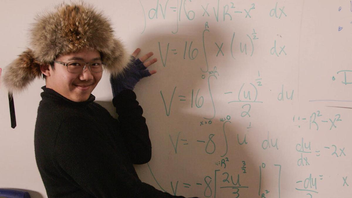 man with furry hat pointing at white board equations