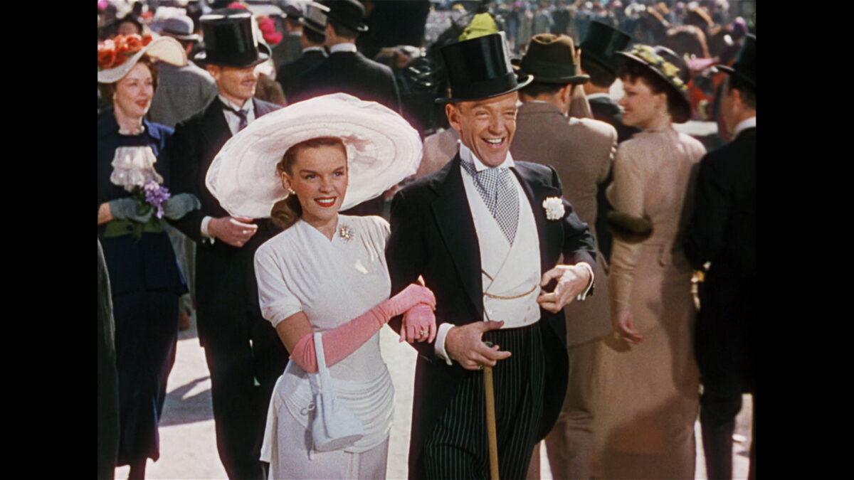 man and woman wearing large hat walking through crowd in formal wear 
