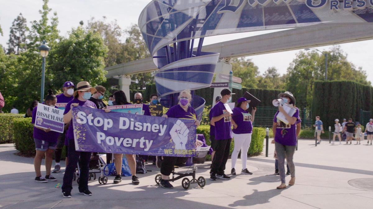 group of people wearing purple shirts and holding sign that says "stop disney poverty"