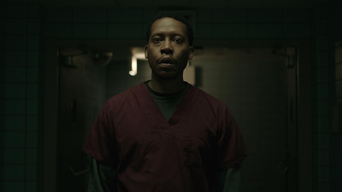 Black man facing forward standing in dark hallway with faint light behind him