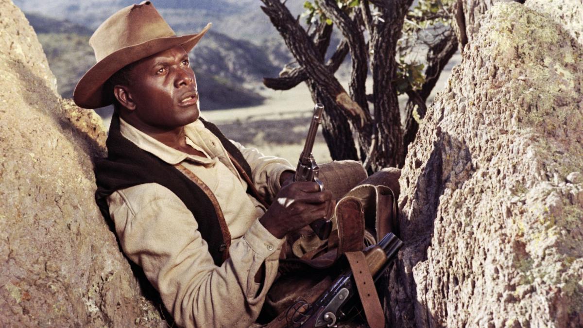 man in cowboy western outfit sitting on rocks with gun in hand