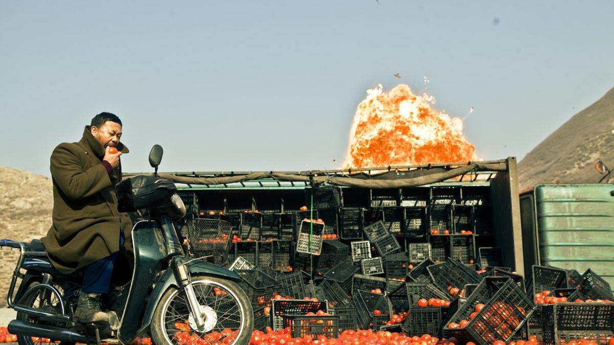 man wearing all black on a motorcycle in front of tomato stand with explosion in backdrop