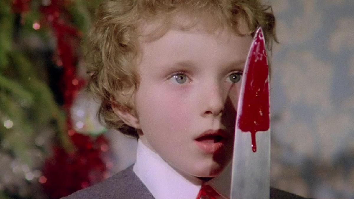 young boy with blond curly hair holding bloody knife in front of face