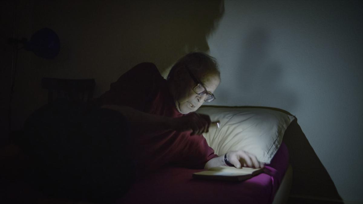 man wearing glasses in bed reading book with flashlight