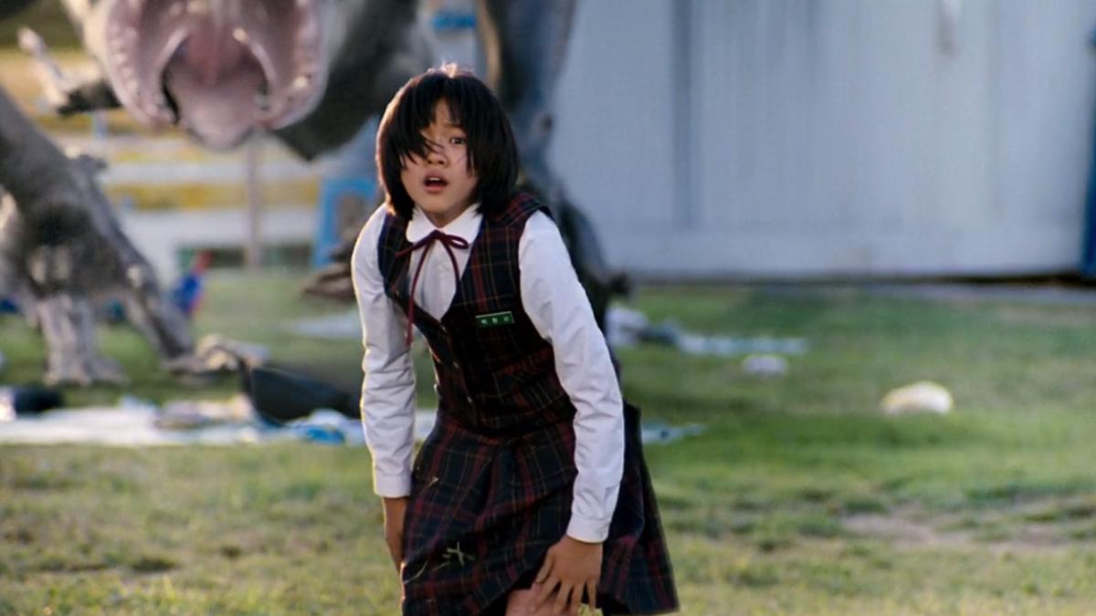 woman in white collared shirt and jumper standing on grass with large monster lurking behind her