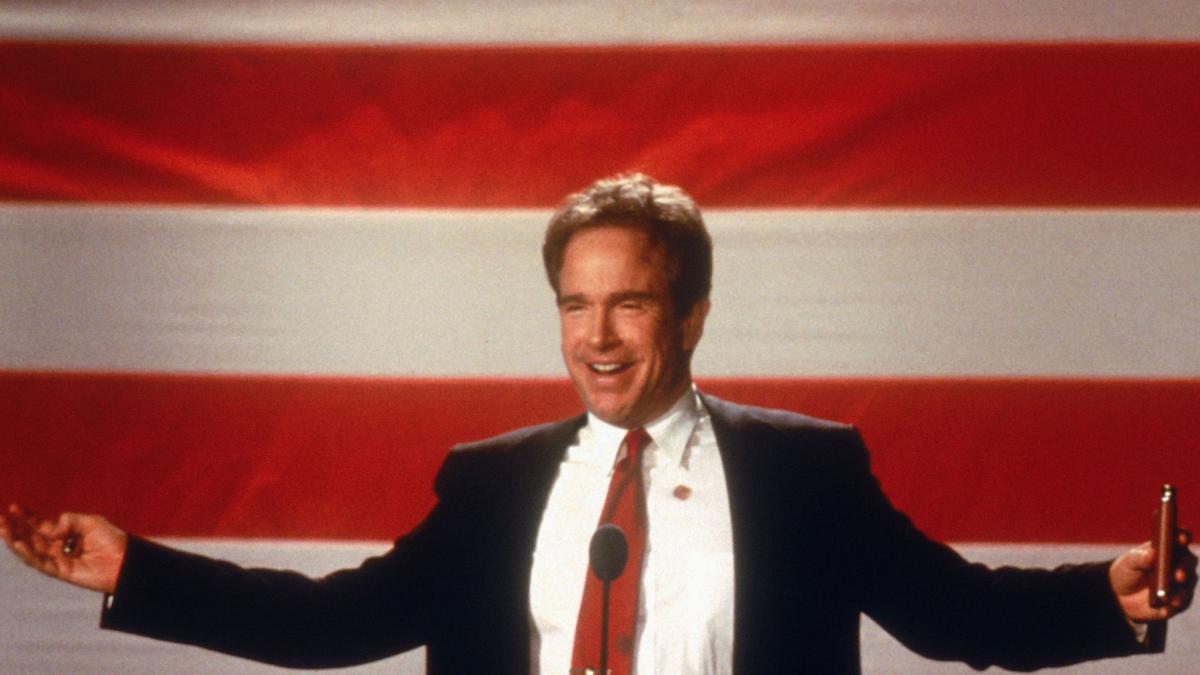 man in suit and red tie standing in front of large American flag with arms outstretched
