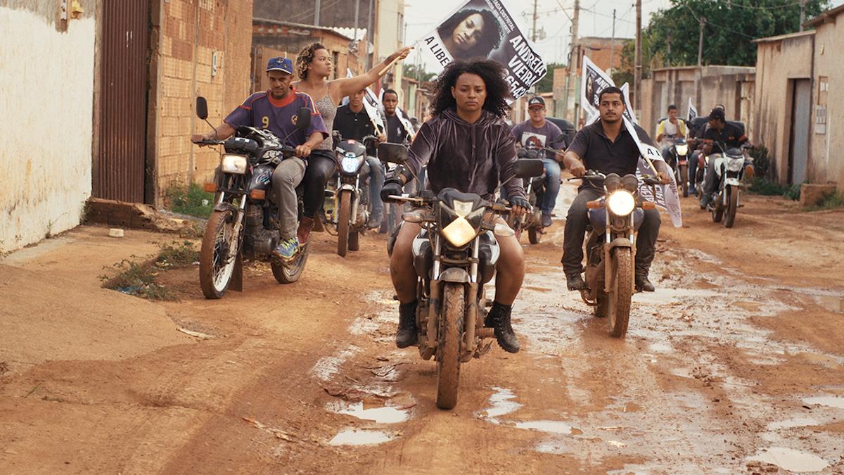 group of people riding down muddy street on motorcycles carrying signs and flags