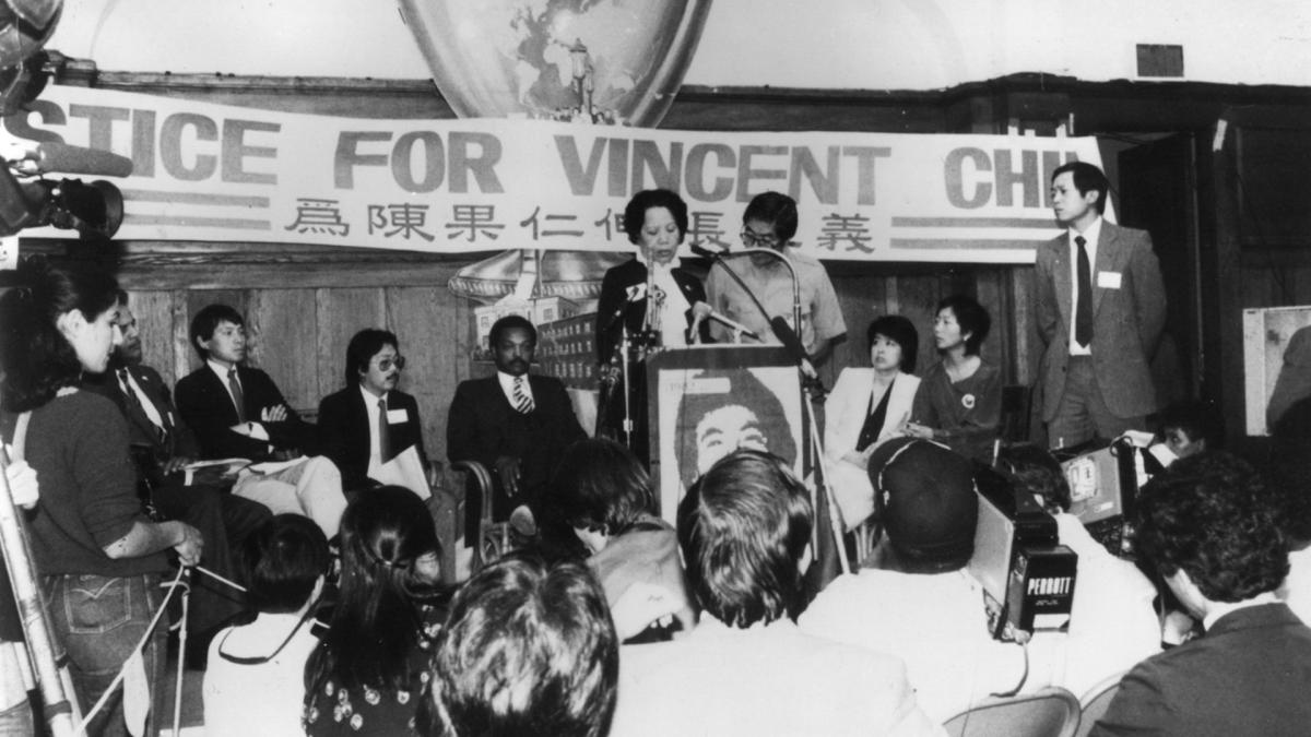 black and white image of woman speaking at podium in front of crowd with signs behind her