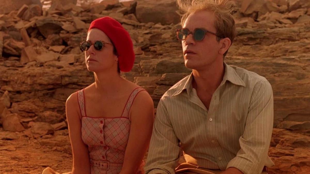 woman wearing red dress, sunglasses, and red beret and man wearing collared shirt and sunglasses sitting next to each other in desert