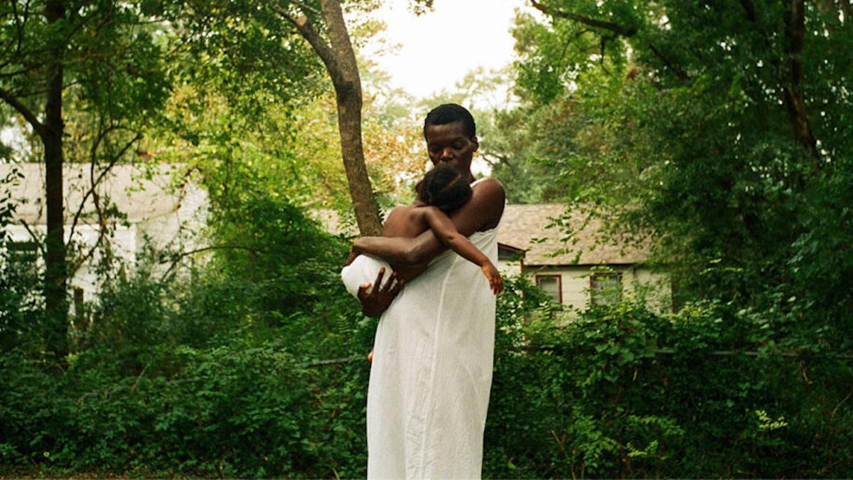 man in white garment holding baby in yard with trees