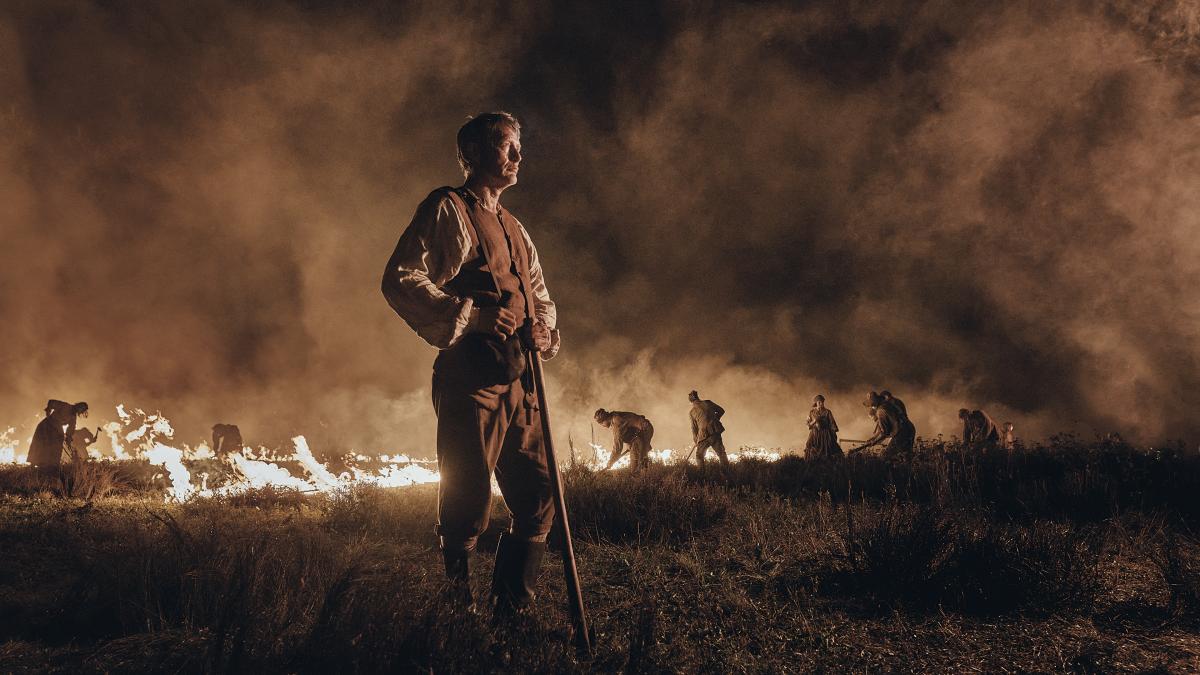 man standing in field with fire in background and men working