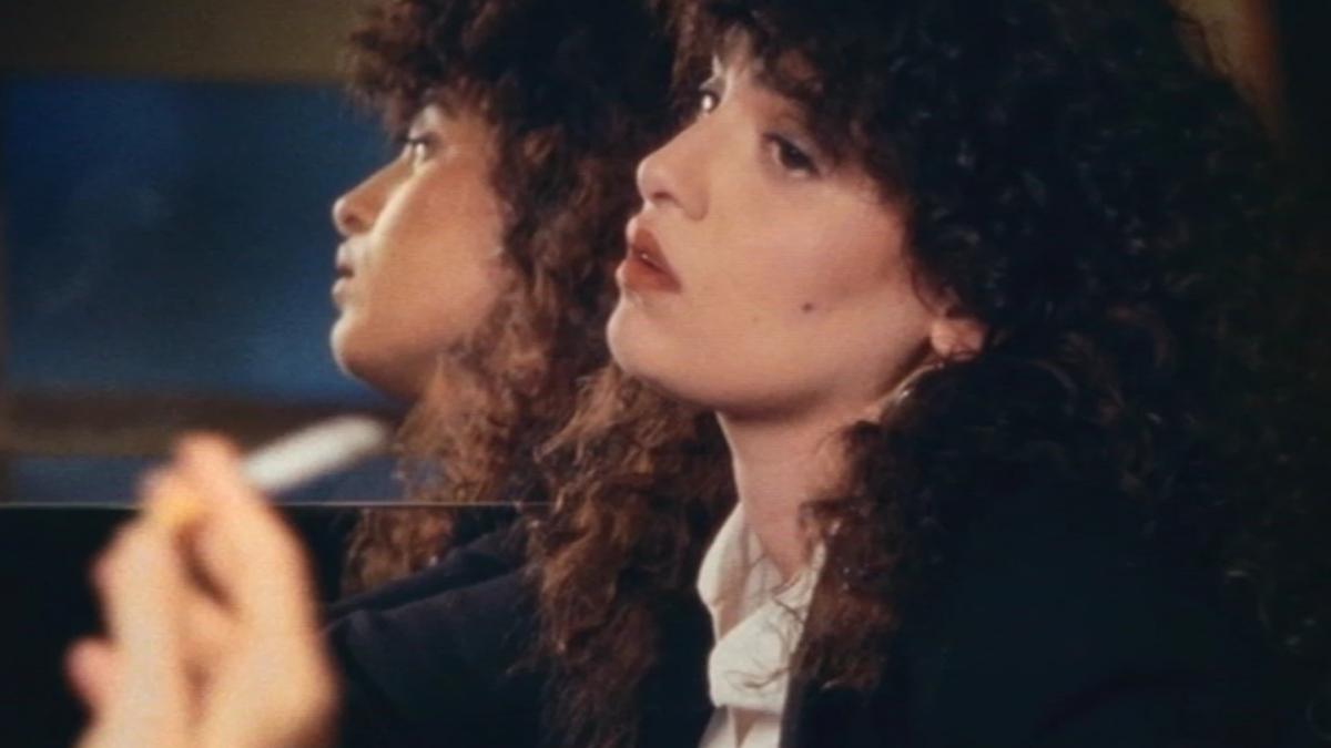 woman wearing white shirt and blazer, smoking cigarette next to mirror
