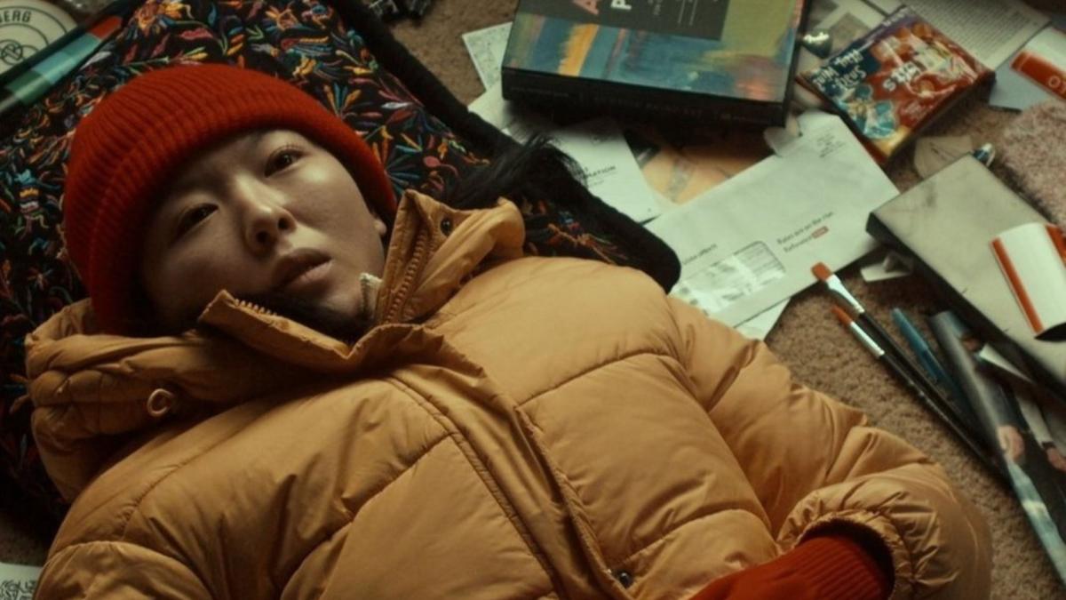 girl in winter coat and hat laying on back on floor near papers and paints