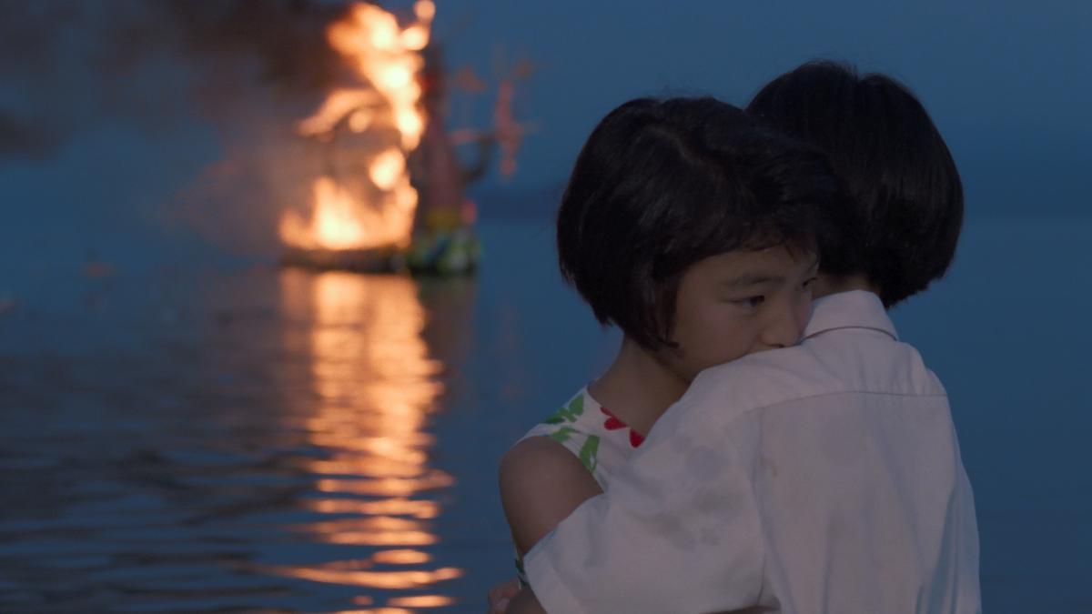 two young children holding each other near water with large fire in background