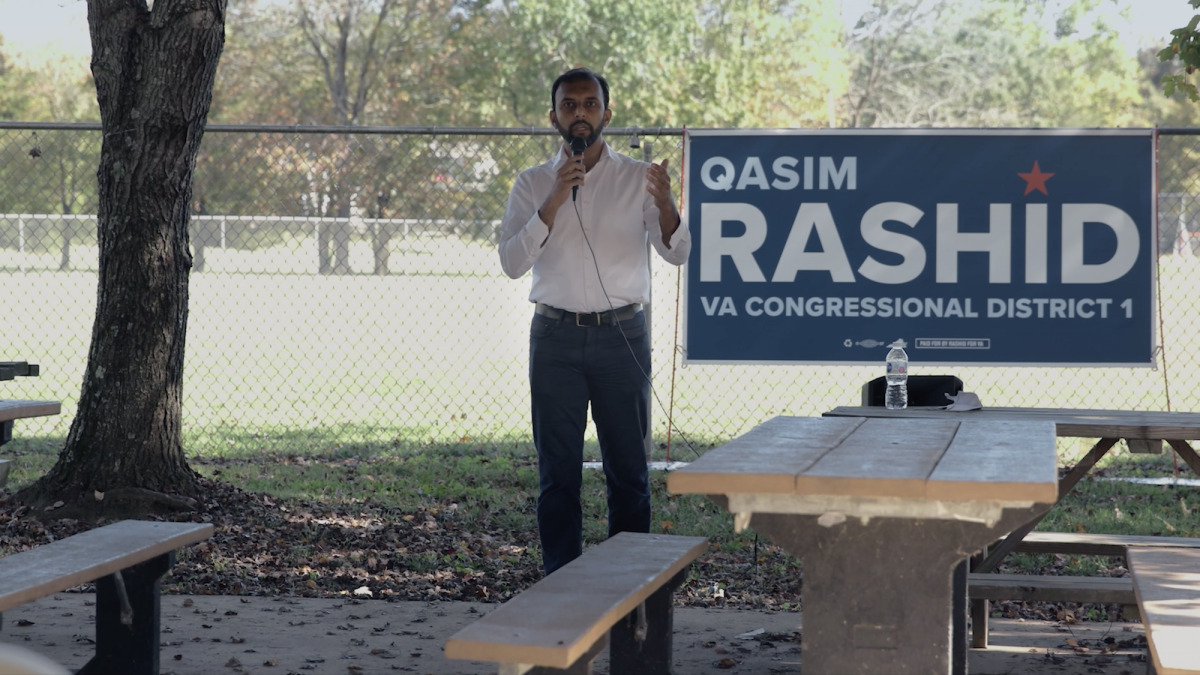 man speaking into mic outside with election sign behind him 