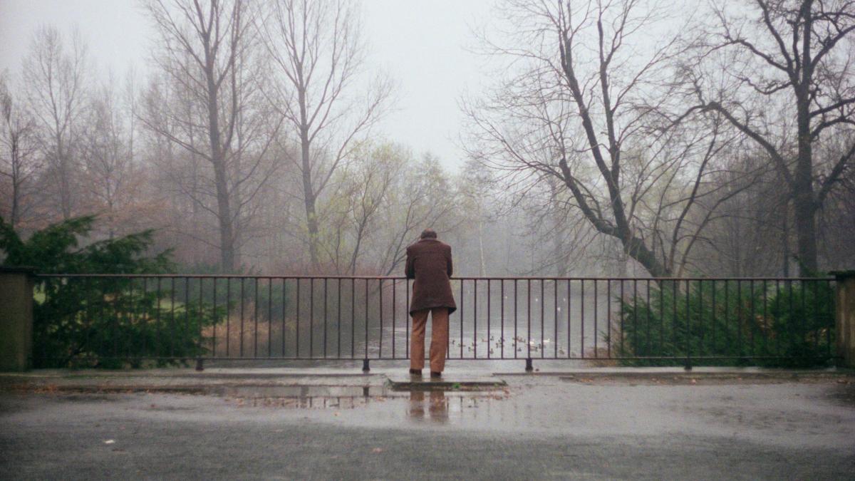 man in brown suit jacket leaning over fence staring down