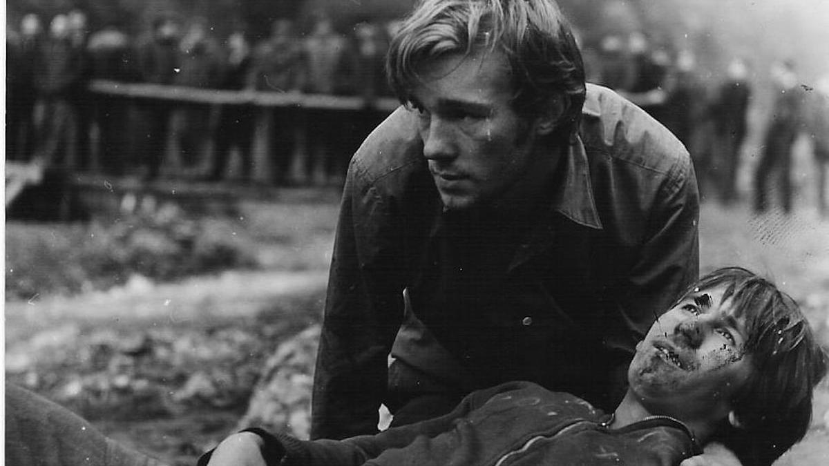 man in muddy field holding dead man up