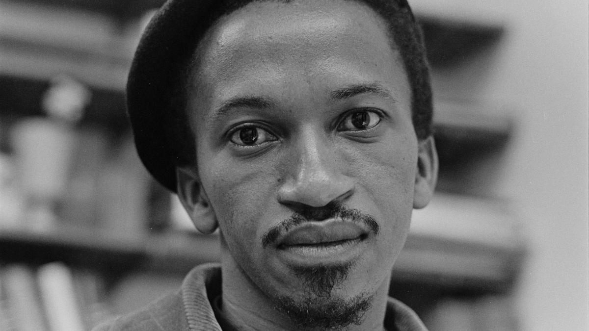 black and white photo of Black man wearing hat in front of bookshelves