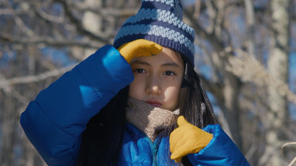 young girl wearing blue winter hat, blue winter coat, and yellow gloves standing outside with trees in background
