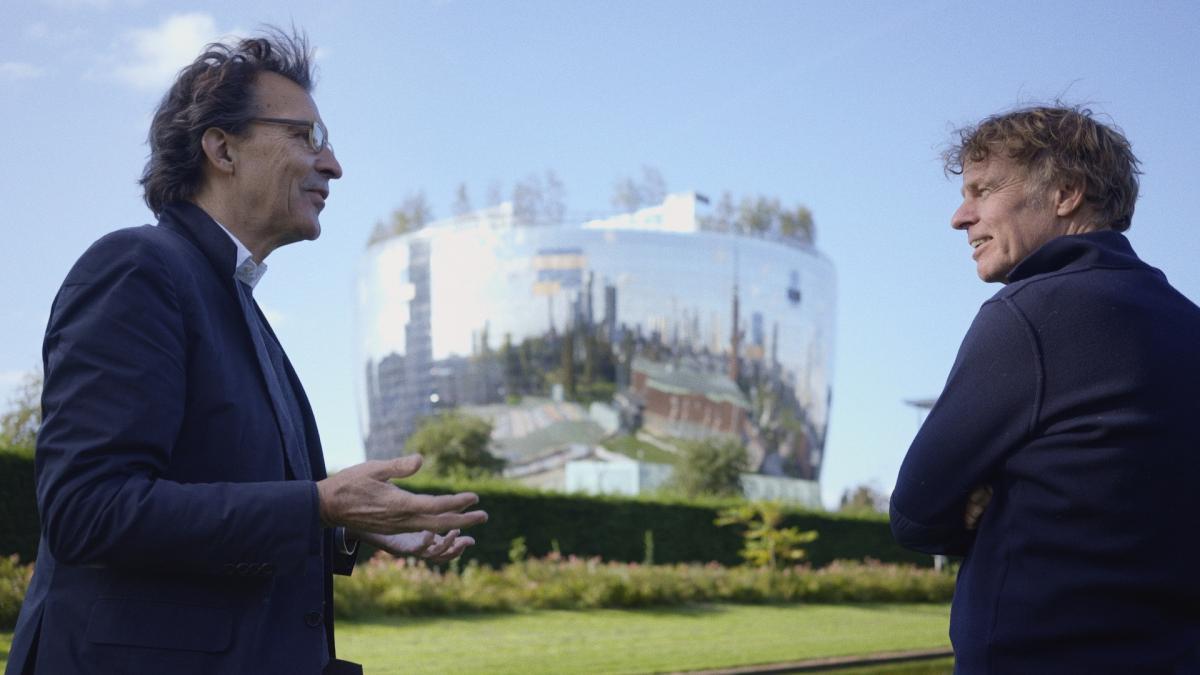 two men in suits standing outside in front of large, silver, futuristic building