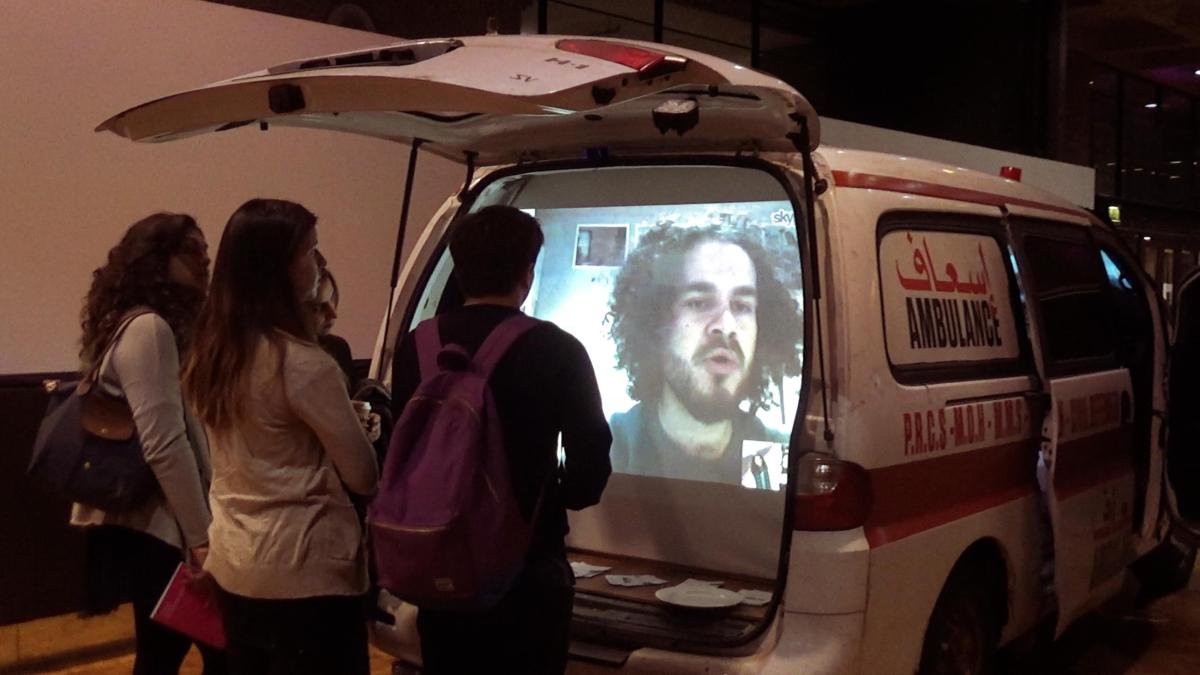 three young people standing behind car with trunk open watching tv screen