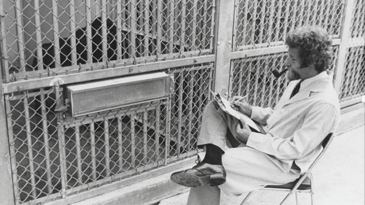 man smoking pipe and writing notes seated in front of cage