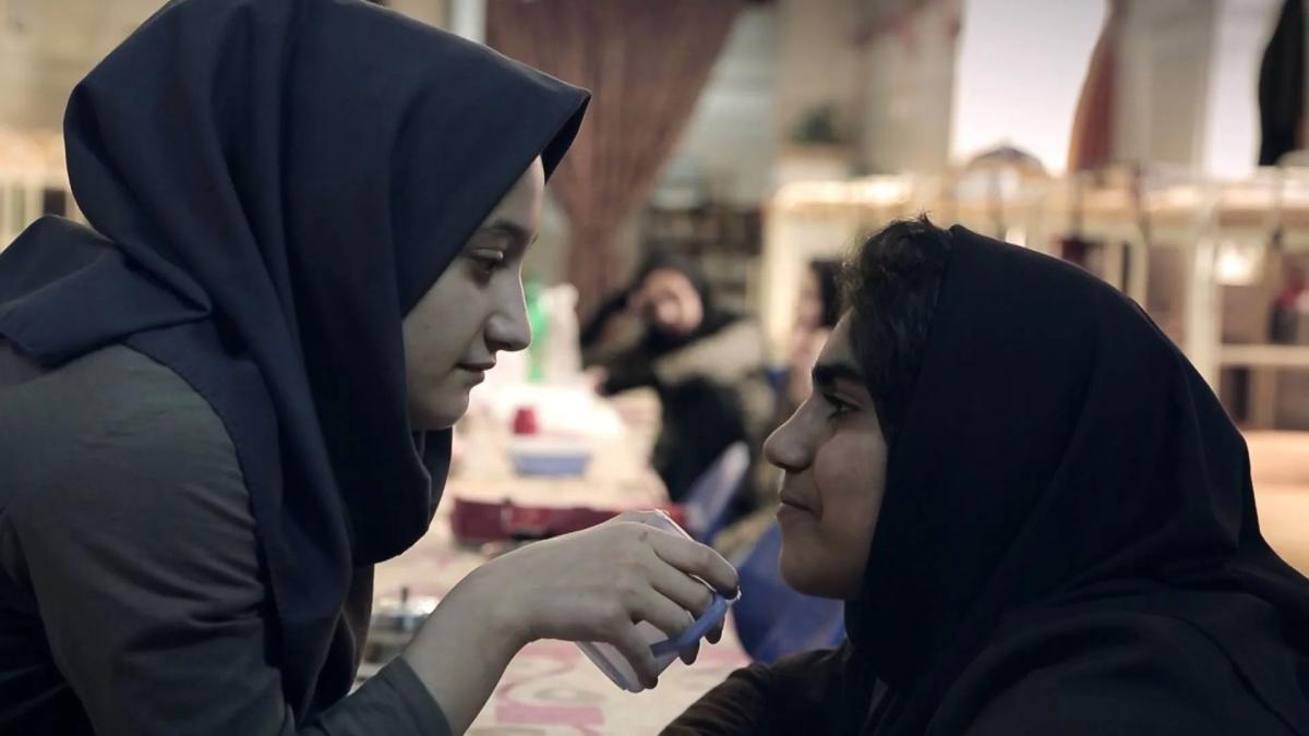 two woman wearing black hijabs sitting outside and looking at each other with one woman's hand on other's chin