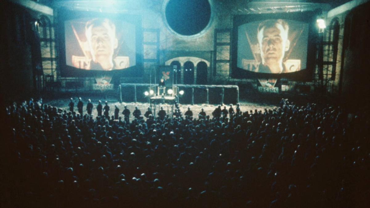 1984, 1984, large crowd facing stage with large monitors of man's face