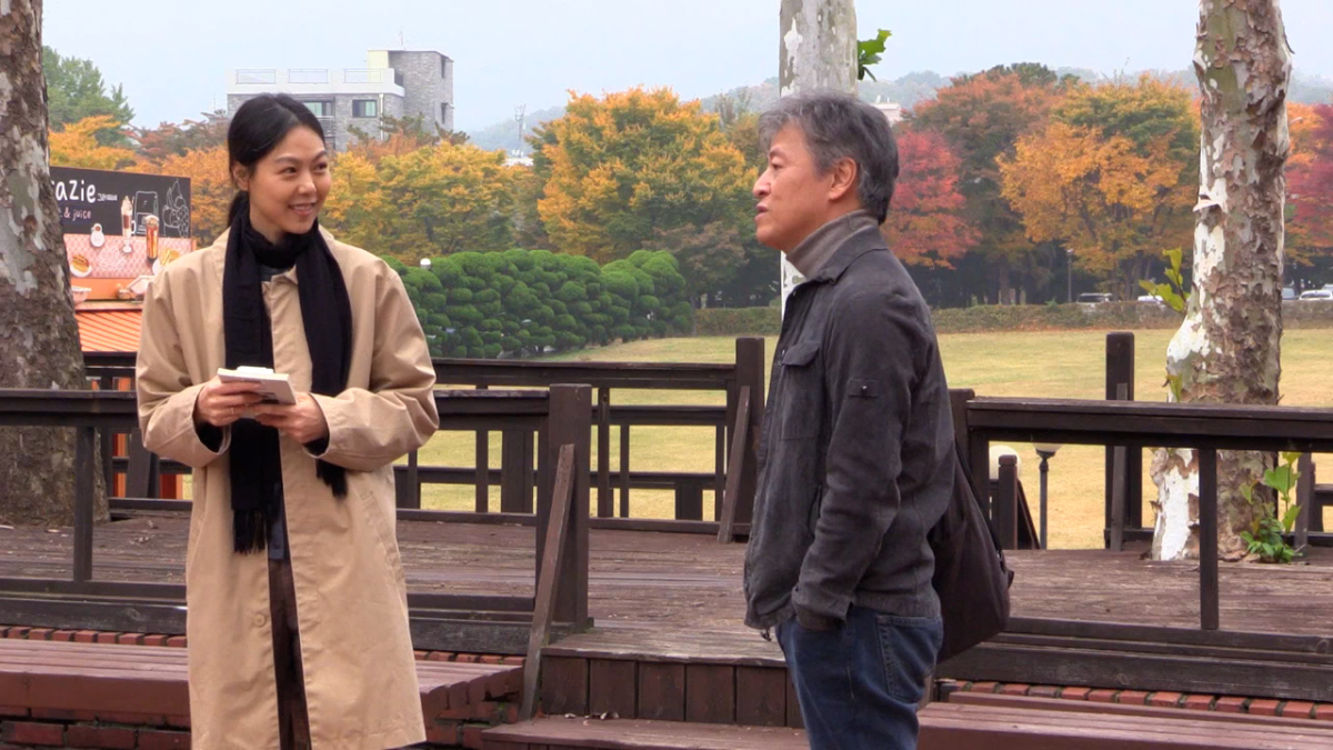 BY THE STREAM, 2024, man and woman in fall coats standing outside on train platform with fall foliage in background