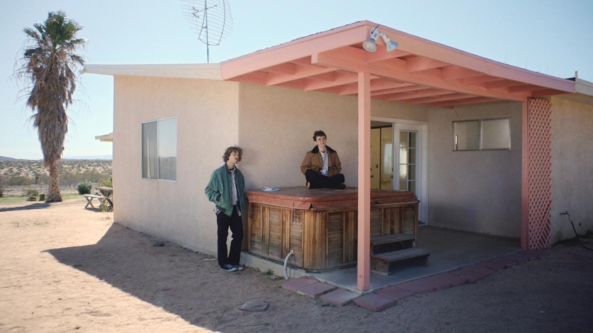 ROOM TEMPERATURE, 2025, two men standing under porch with hands in pocket of a white stucco house with pink roofing