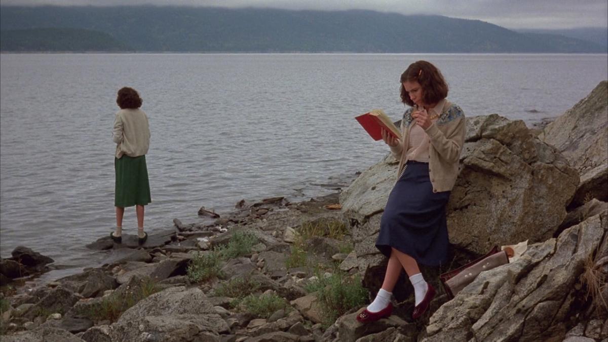 HOUSEKEEPING, 1987, two girls wearing sweaters and skirts standing on rocks by lake with one reading a book