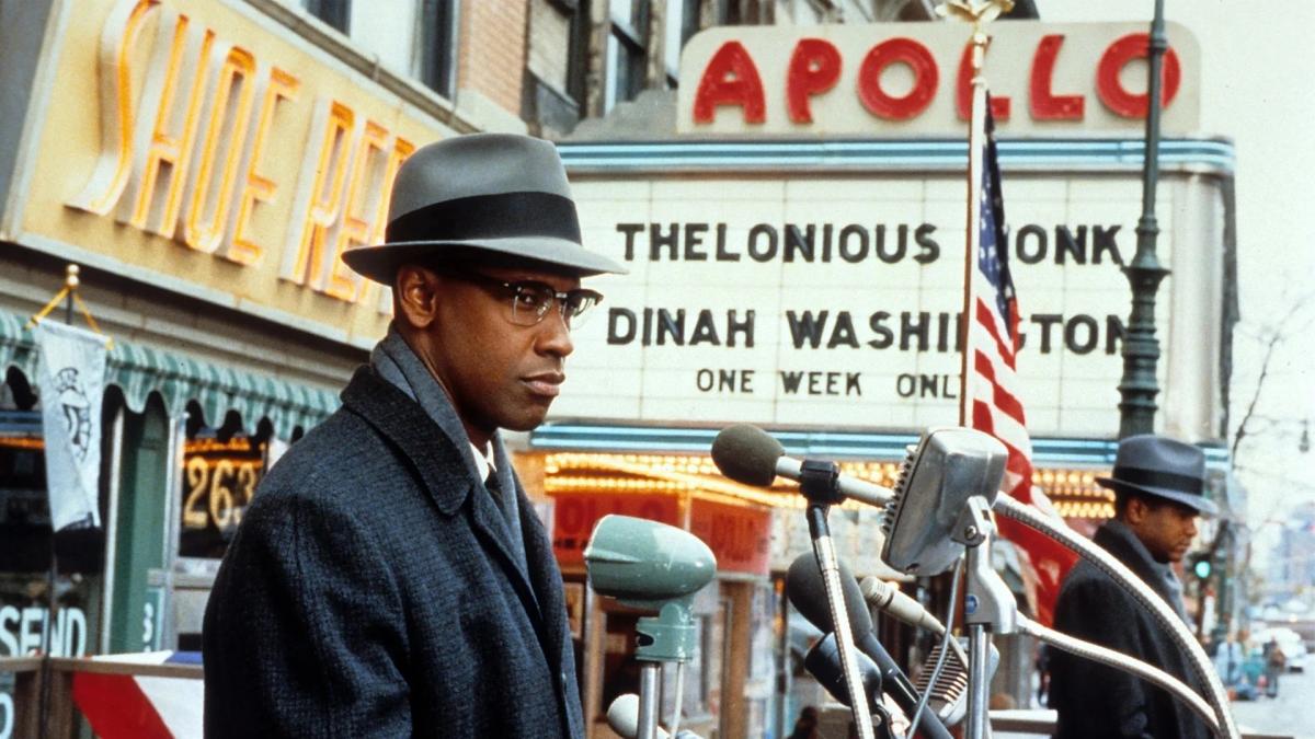 MALCOLM X, 1992, man in hat and jacket standing in front of podium outside on city street