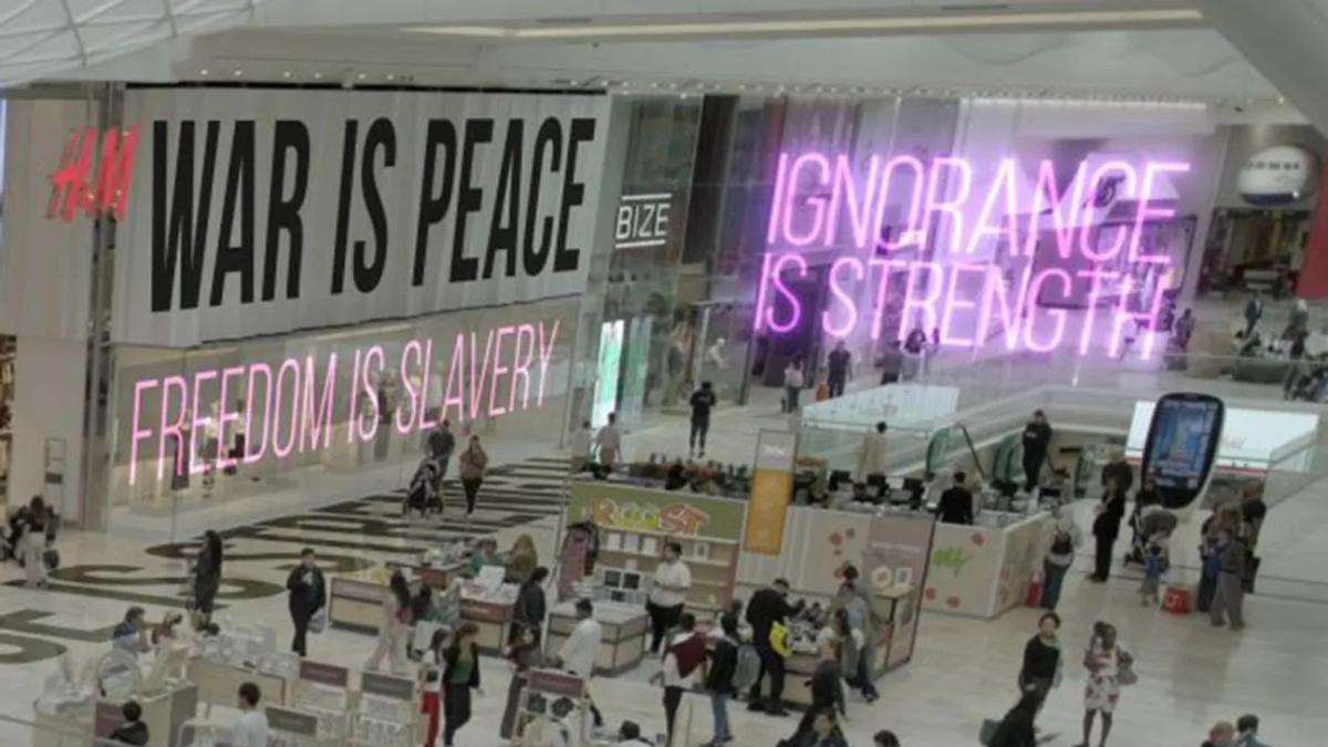 ORWELL: 2+2 = 5, 2025, shopping mall from above with signage overlaid that says "War is Peace," "Ignorance is Strength," and "Freedom is Slavery"