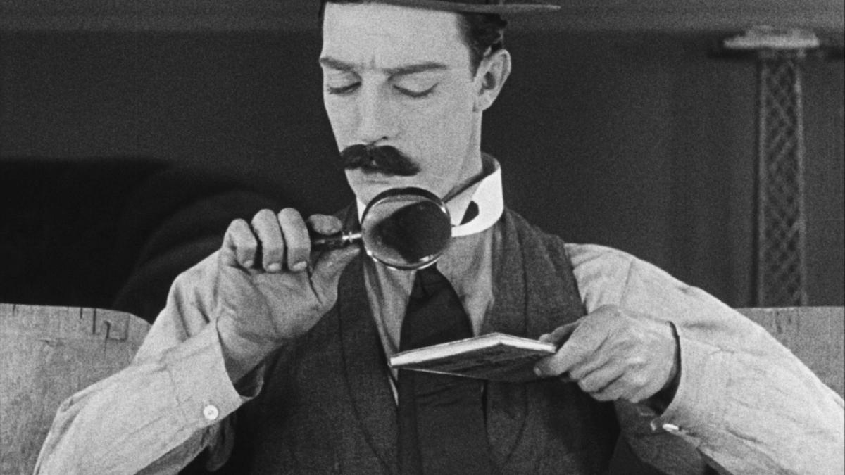 SHERLOCK JR., black and white image of man in hat and vest inspecting item closely with magnifying glass
