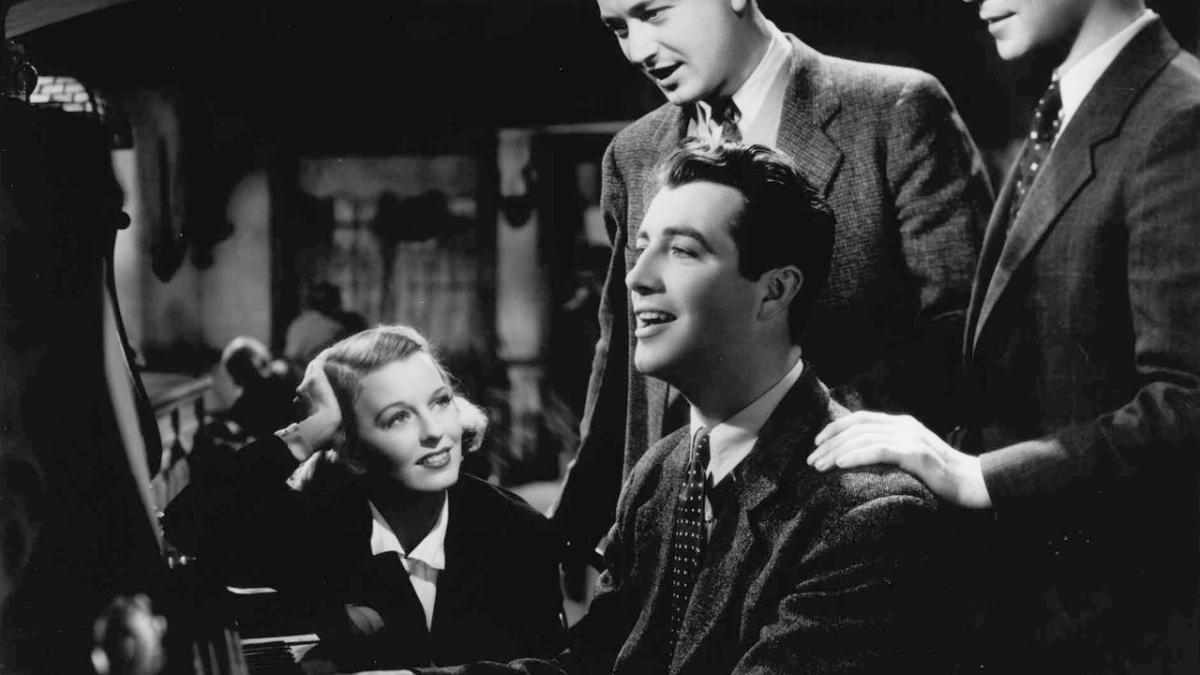 THREE COMRADES, 1938, black and white image of man playing piano with woman sitting next to him and two men standing behind him with hands on shoulders