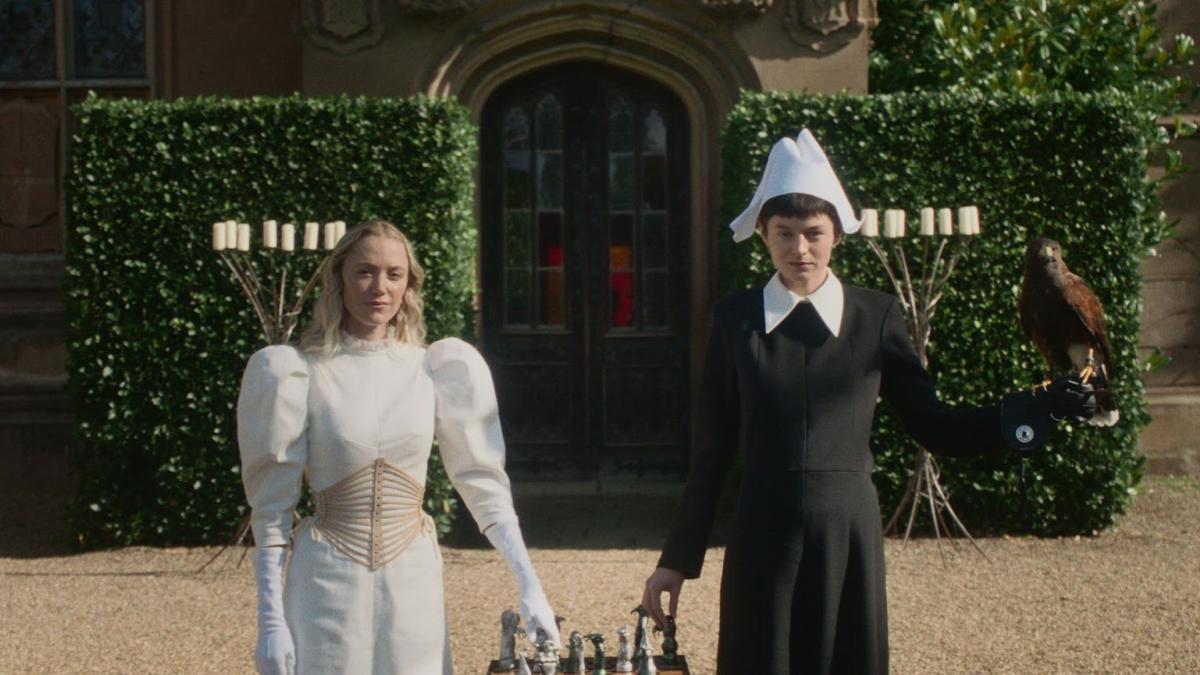 100 NIGHTS OF HERO, 2025, woman in white dress and person in black dress and cap standing holding basket between them walking away from bushes