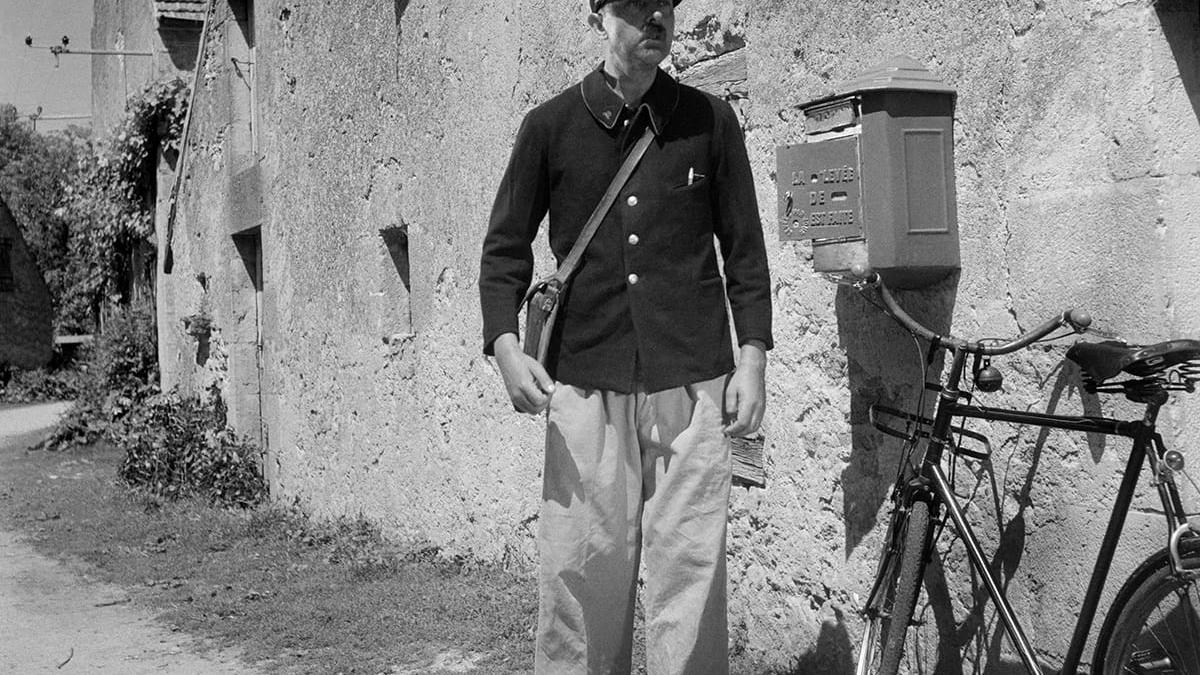 black and white image of man in mail carrier uniform standing near building and bicycle 